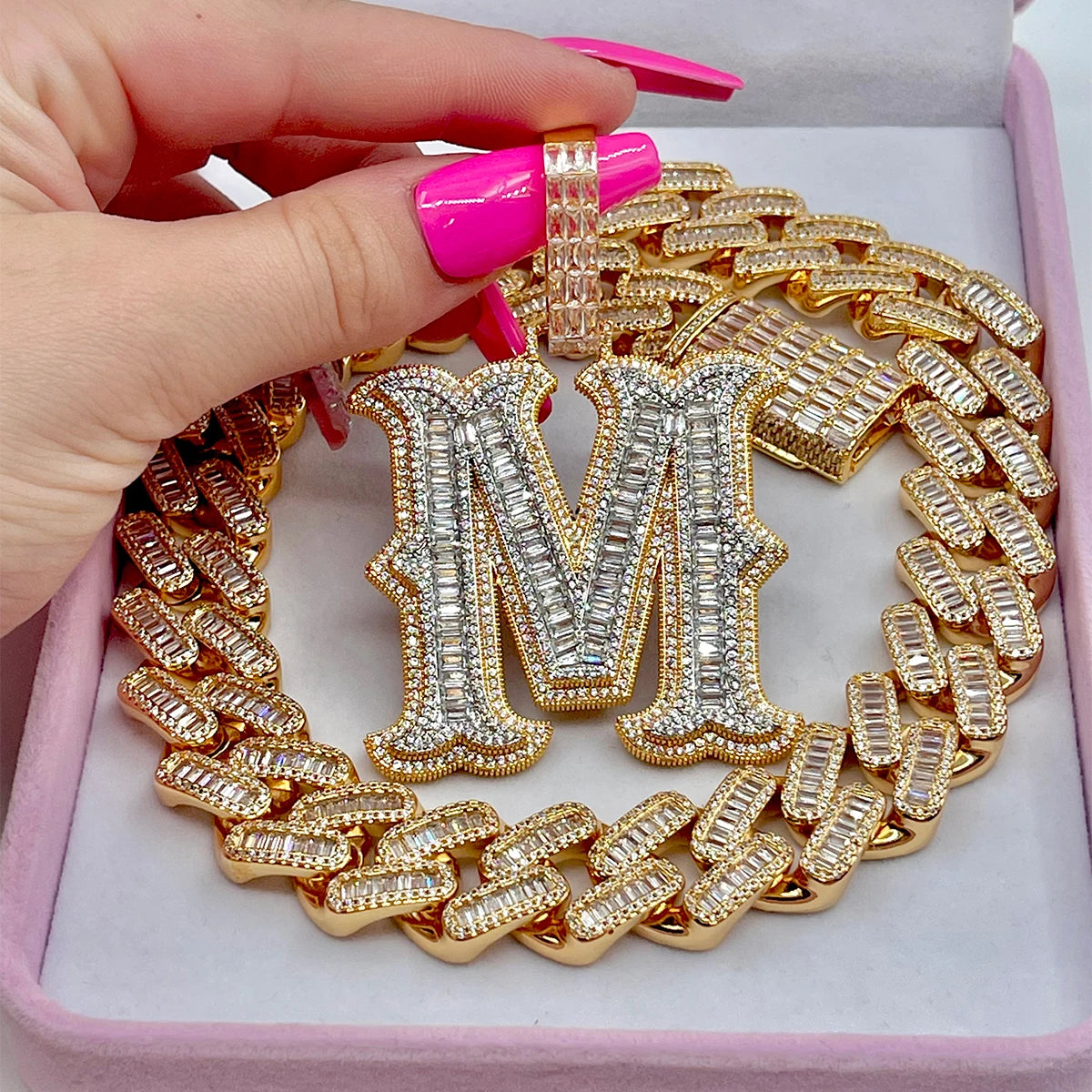 Gold chain necklace with a large letter 'M' pendant held by a hand with pink nail polish.