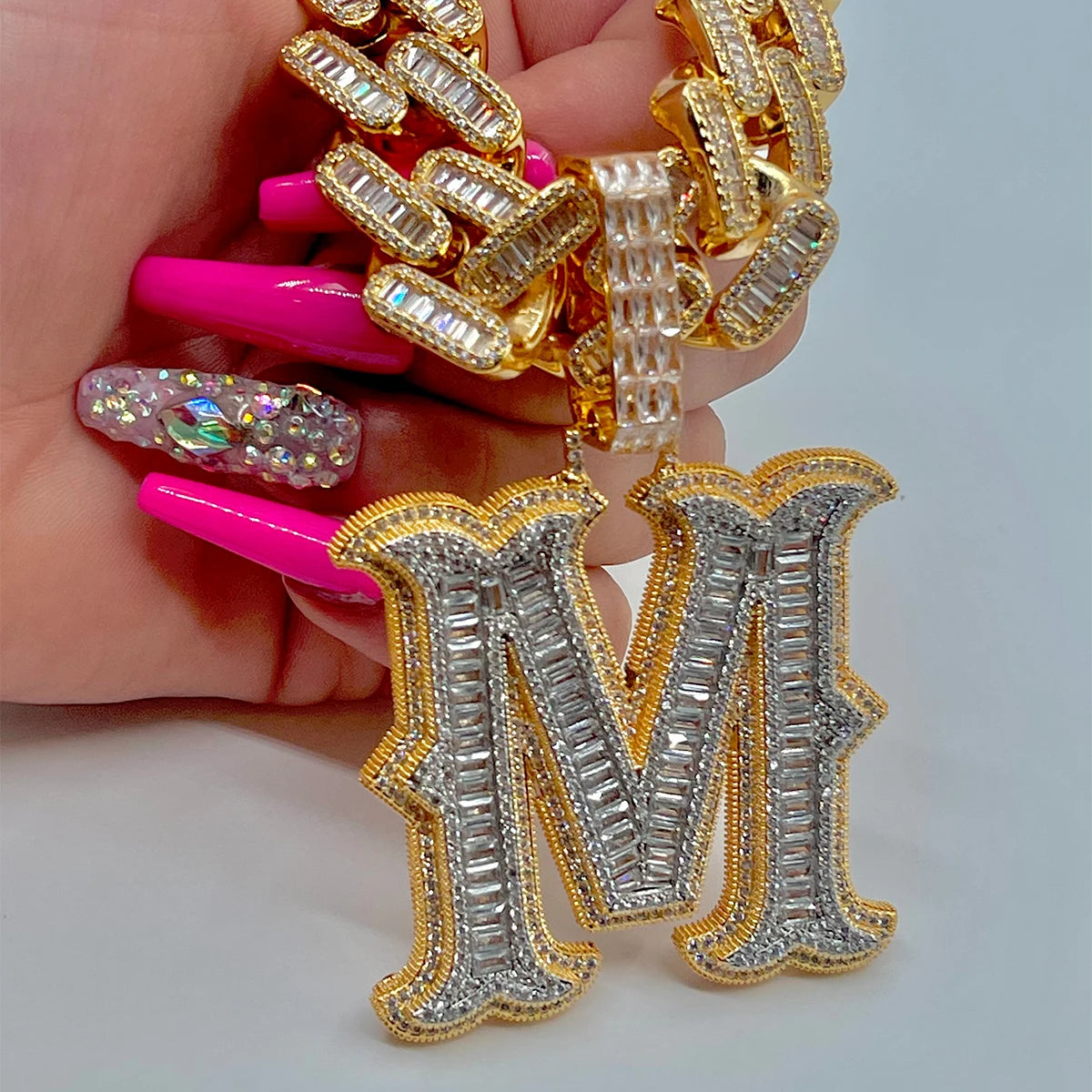 Gold and silver letter 'M' earrings held by a hand with pink nail polish.
